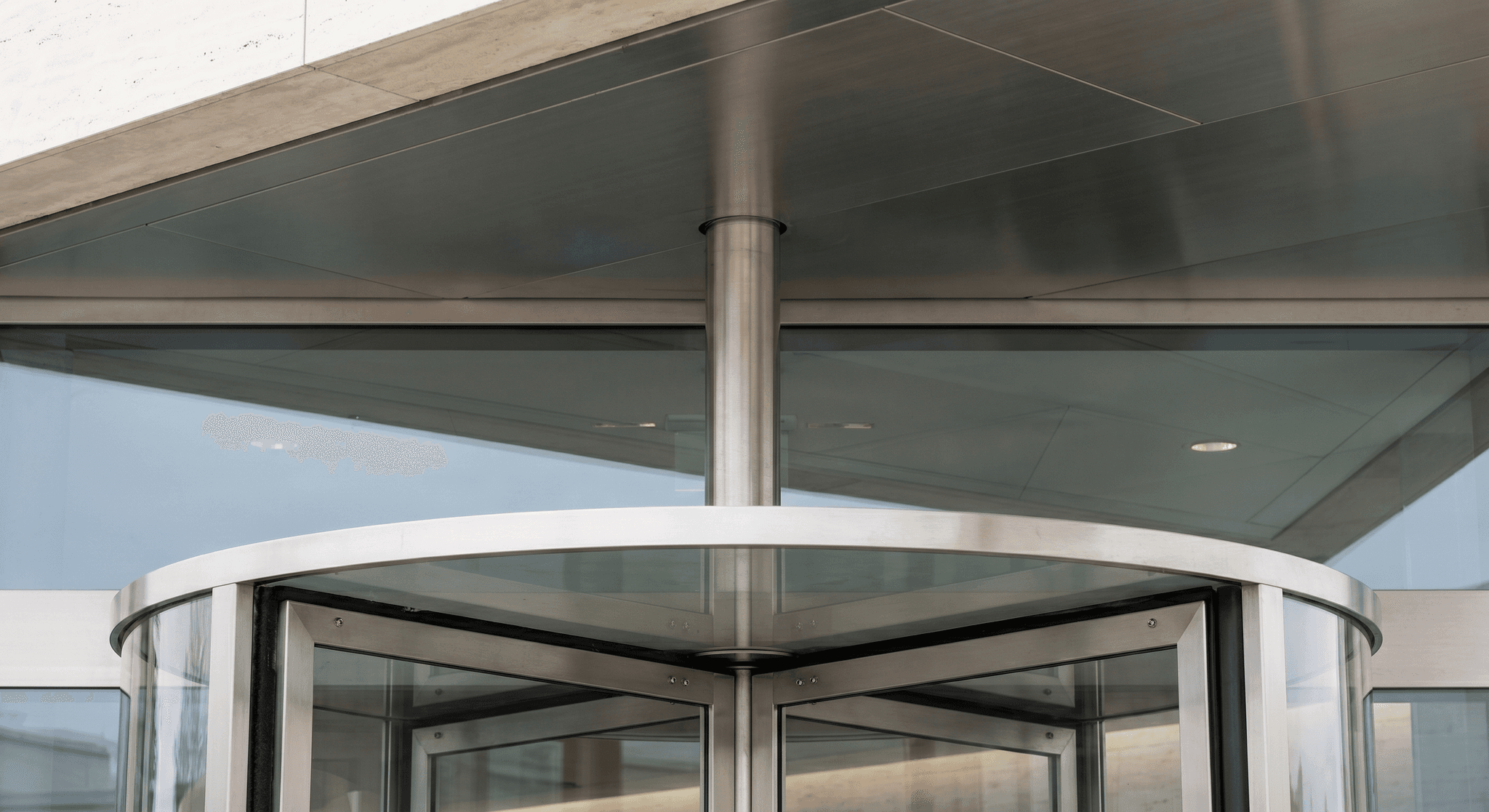 Detail of revolving door crown housing, vertical brushed stainless steel pivot axis, machined bushing, recessed travertine lintel