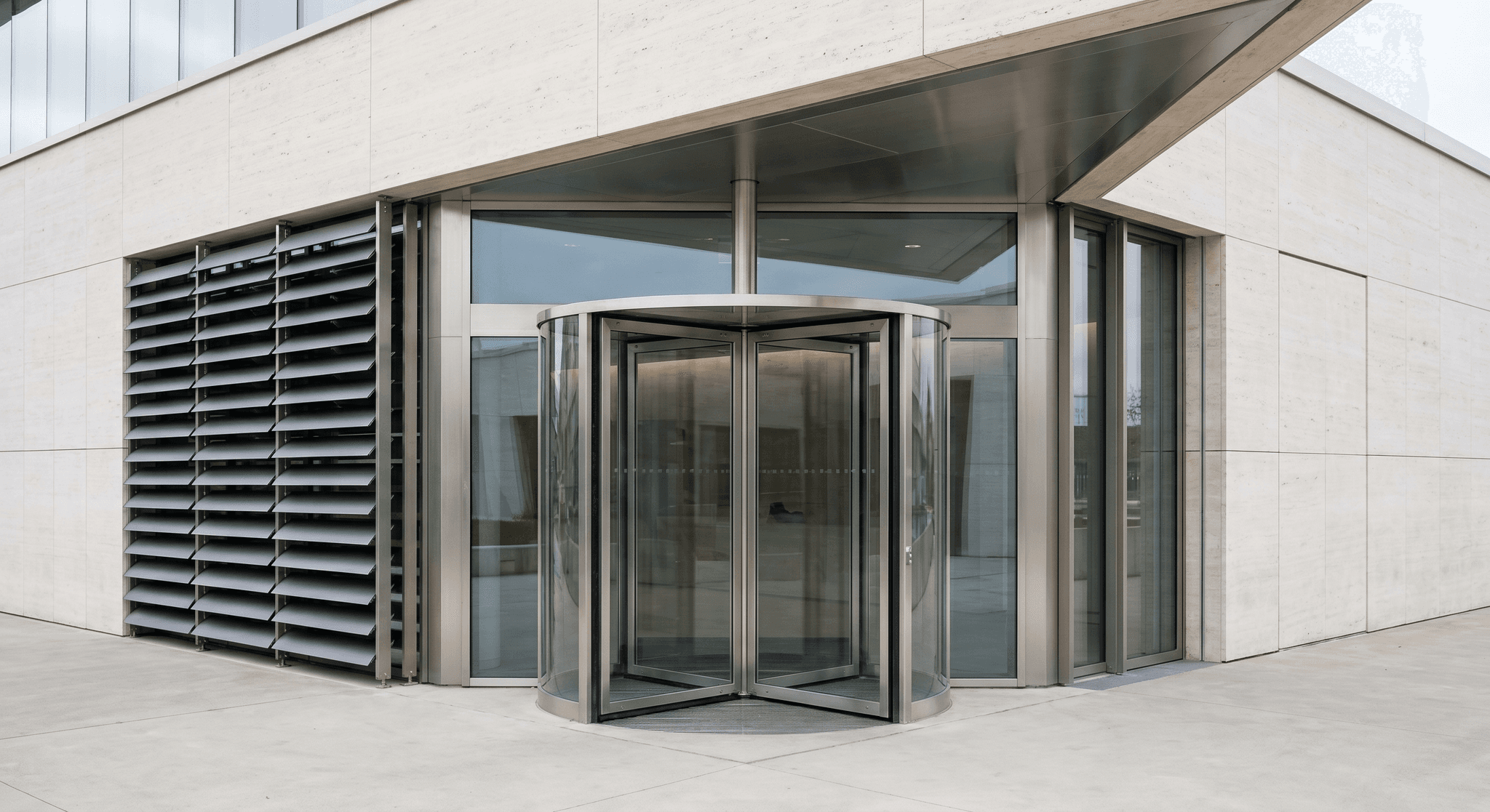 Signature institutional entrance, four-leaf revolving door in glass and stainless steel, kinetic facade with motorised louvres, lateral motorised sliding door, polished concrete plaza